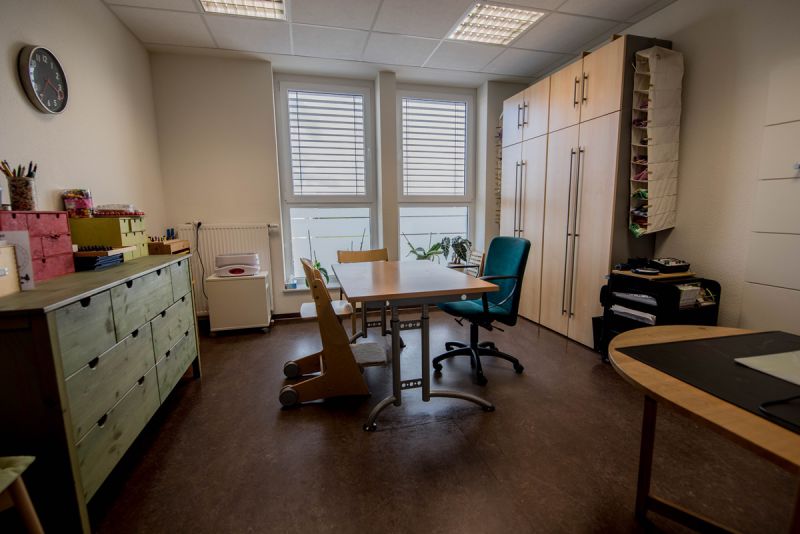 Therapieraum 1 - Ein Tisch mit zwei Stühlen in der Mitte, ein Regal auf der lin ken Raumseite und im Hintergrund rechts ein großer Schrank