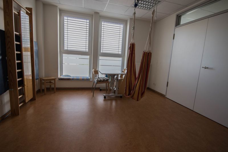 Therapieraum 2 - hinten rechts steht ein Tisch mit zwei Stühlen, im Vordergrund ist eine Schlaufenschaukel zu sehen, an der linken Wand ist eine Sprossenwand befestigt