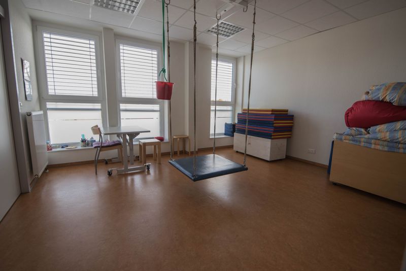 Ein Blick in den Terapieraum 3, man sieht links einen Tisch mit zwei  Stühlen, daneben eine Matte auf dem Boden mit Stangen an den Ecken, die vom Boden zur Decke reichen